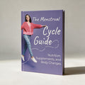 A hardcover book titled "The Menstrual Cycle Guide: Nutrition, Supplements, and Body Changes" with a purple cover, standing upright on a light gray surface against a neutral background. The cover features a photo of a woman in a pink patterned cardigan and blue jeans.